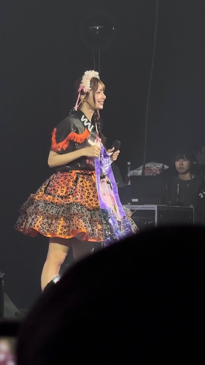 A young woman performing on a dimly lit stage, wearing a black fringed top with red tassels, a colorful animal-print ruffled skirt, and a white flower headband, holding a microphone and a purple cloth.