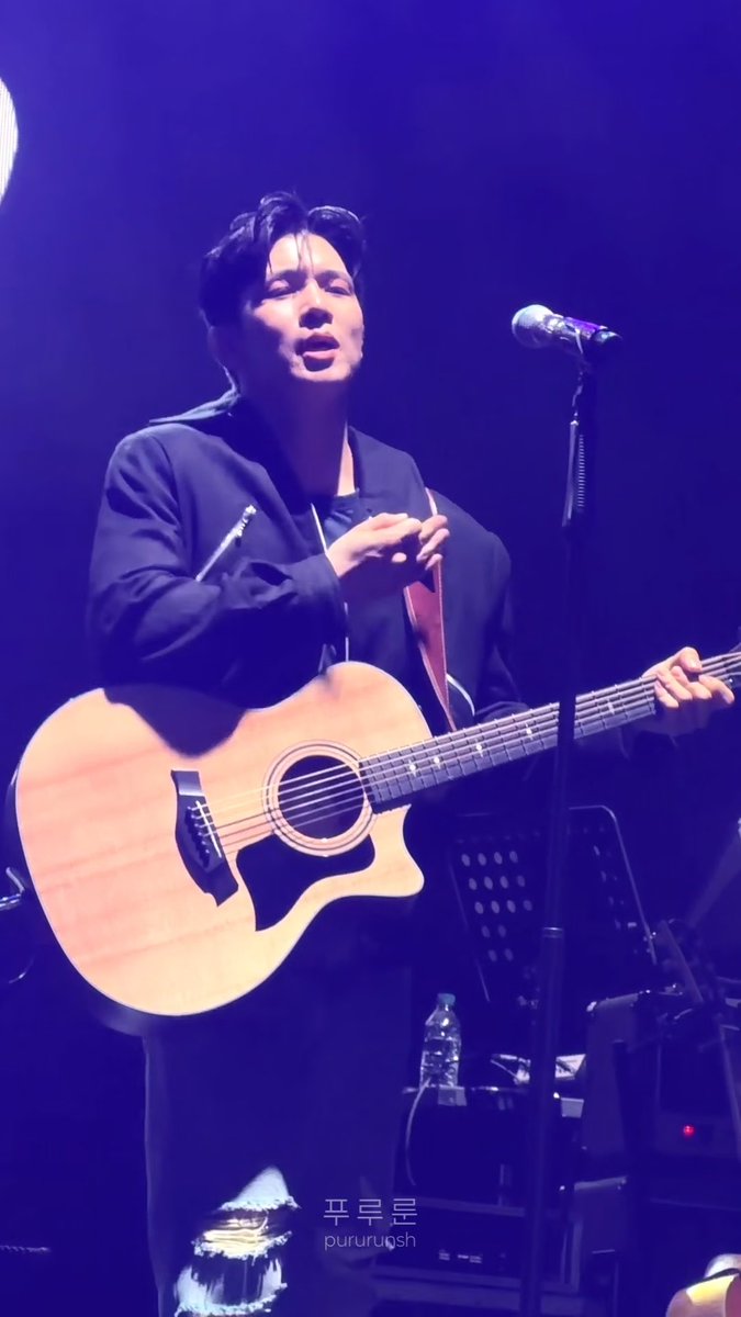A man with dark hair performs on stage playing an acoustic guitar and singing into a microphone, wearing a dark jacket, with a watermark reading '平早早 pururu9sh' visible.