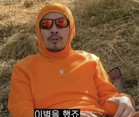 A man wearing an orange hoodie and sunglasses sits relaxed against a haystack, with Korean subtitles reading "이번을 해주" ("doing this time") at the bottom, matching the laid-back, humorous tone of the caption about Noh Hong-chul's casual YouTube reveal about his past relationship with a female doctor.