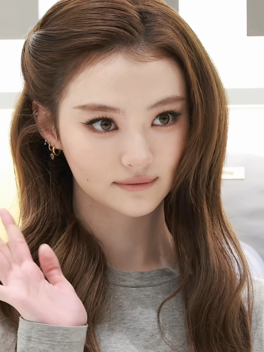 A close-up portrait of a young woman with long brown hair, wearing a gray shirt and gold earrings, who appears to be waving.
