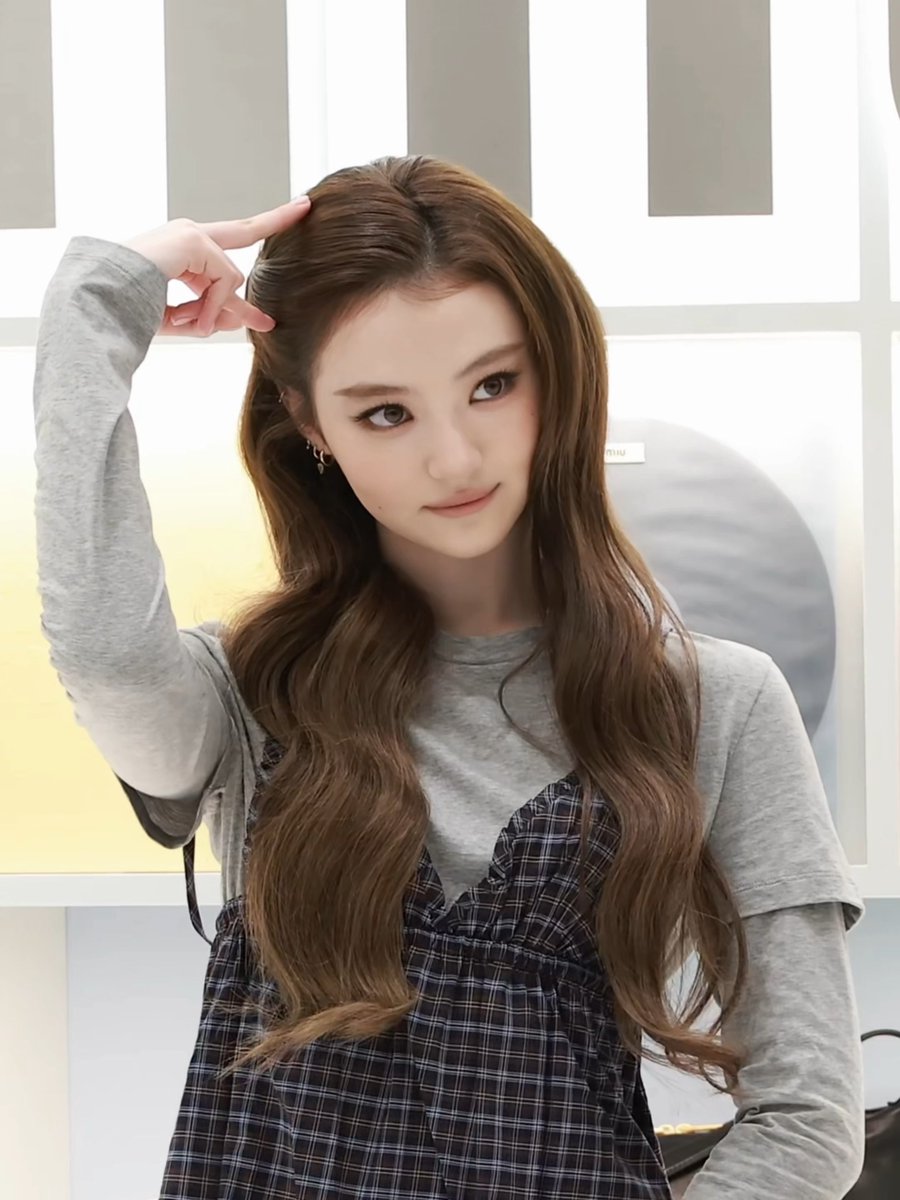 A young woman with long wavy brown hair posing indoors, wearing a gray long-sleeve top under a black and white plaid pinafore dress, with one hand raised near her head and a slight smile.