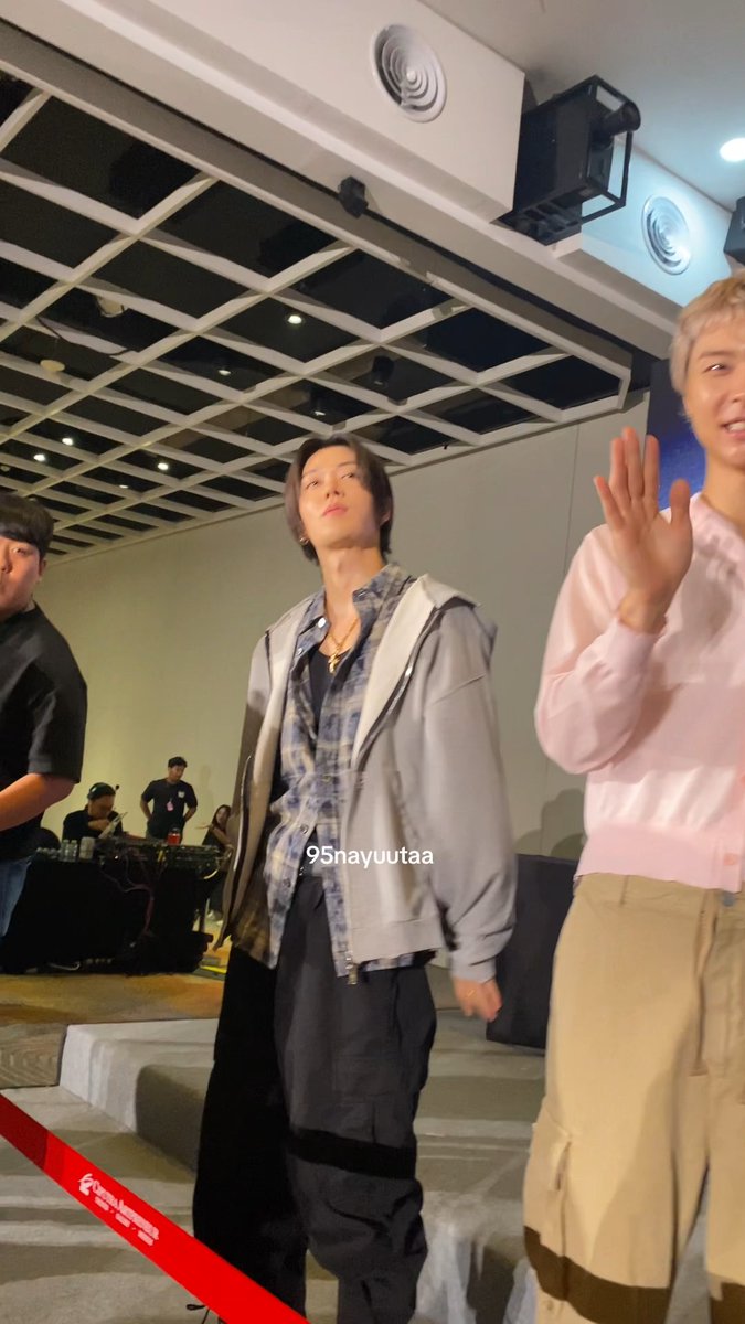 The image shows NCT127's Yuta posing at what appears to be a fan event, wearing a gray hoodie layered over a plaid shirt with dark cargo pants, while a smiling friend in a pink sweatshirt waves beside him. The watermark "95nayuutaa" is visible, and the caption "HALO MY YUTA AND FRIEND#NCT127" reflects the excited fan greeting toward the idol and his companion.