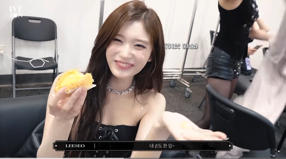 Leeseo from IVE winks while holding a piece of yellow pastry in what appears to be a behind-the-scenes vlog. Korean text on screen reads '(히힛 떠출)' and 'LEESEO | 네 손도 한입~'.