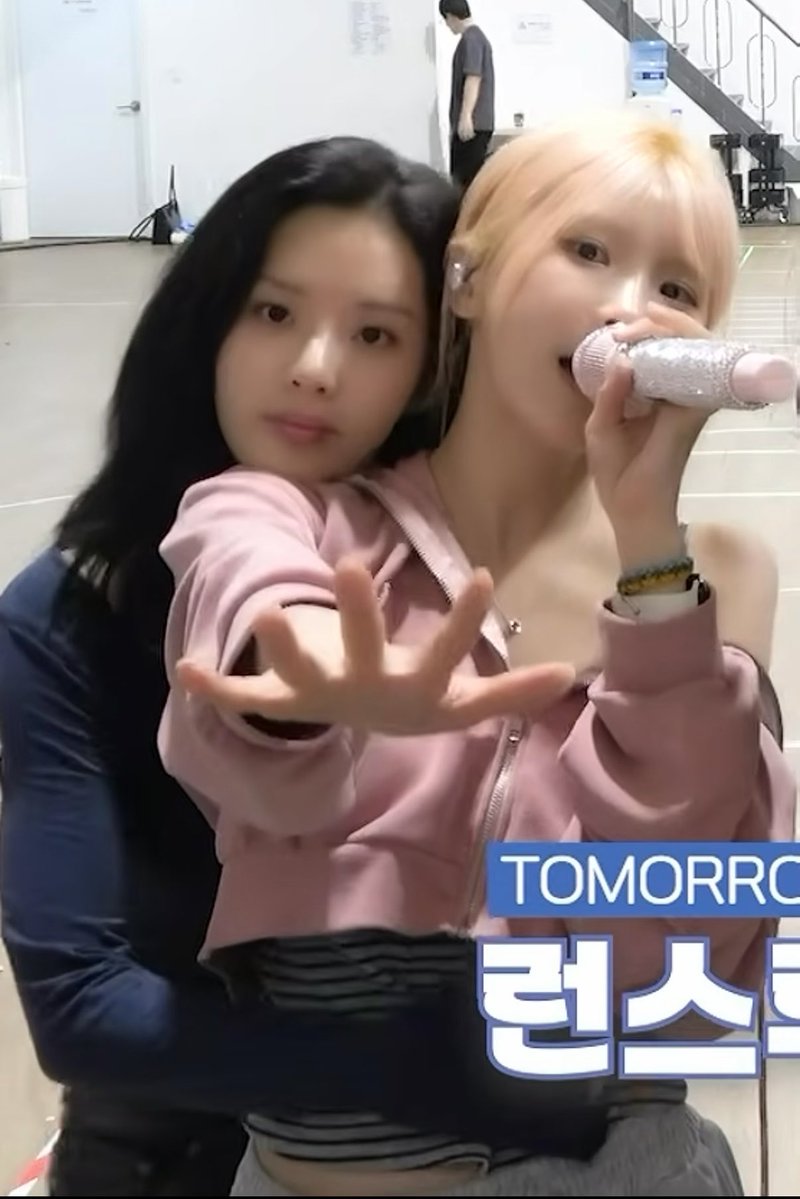 Two women, one with long dark hair and the other with blonde hair holding a pink microphone, pose together in what appears to be a backstage area with "TOMORRO" and Korean text "런스" visible.