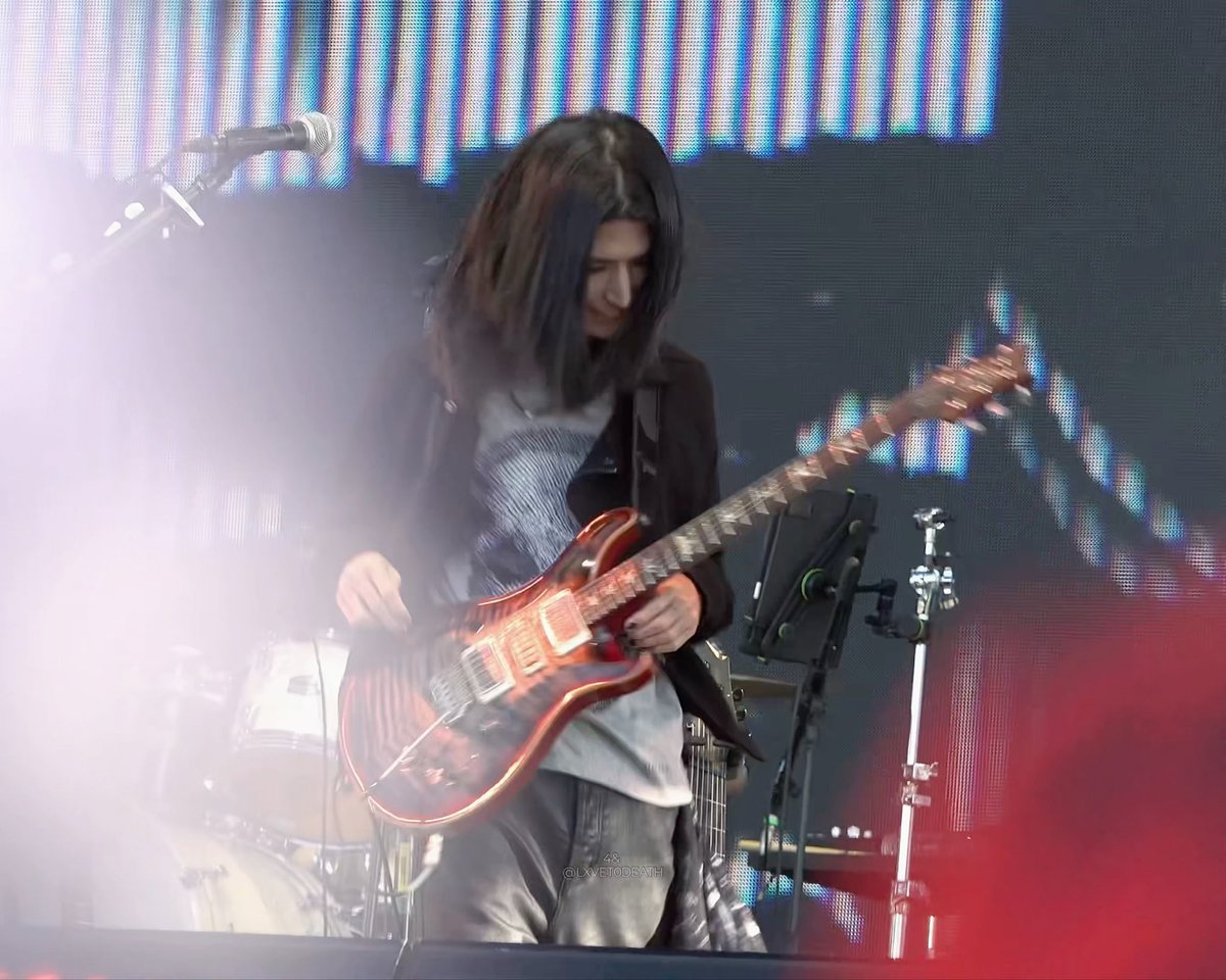 A musician with long dark hair performing on stage, playing a red PRS-style electric guitar under bright lights and smoke effects.