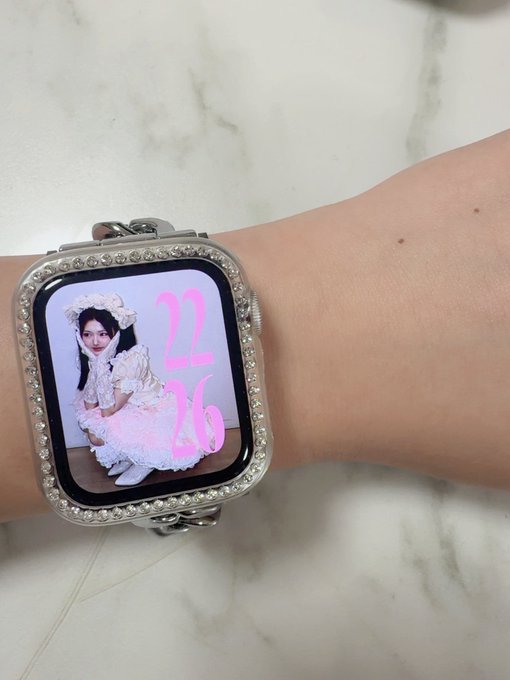 A smartwatch with a crystal-encrusted band displays a pink watch face showing '12:16' alongside a photo of a young woman wearing a white floral dress and headpiece.