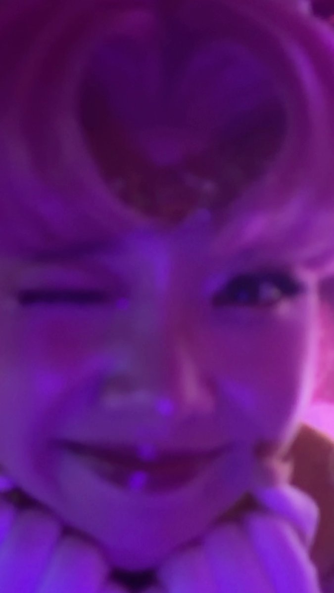 A blurry, purple-tinted photo of a smiling person winking and making a hand gesture near their face.