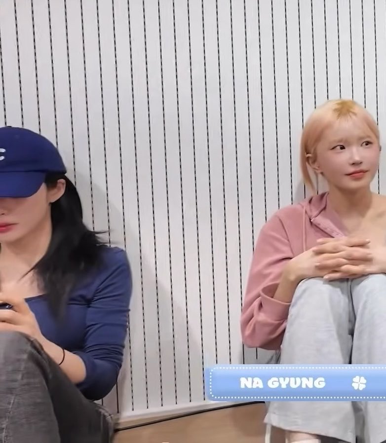 Two women sit against a white striped wall, with the blonde woman on the right (labeled "NA GYUNG") wearing a pink hoodie and gray sweatpants, while the other woman on the left wears a navy blue cap and top, looking down at her phone. The image, paired with the caption "Ahhh, don't do it, don't do it ..", suggests Na Gyung is playfully reacting to something dramatic happening off-camera, likely teasing or warning someone in a lighthearted way.