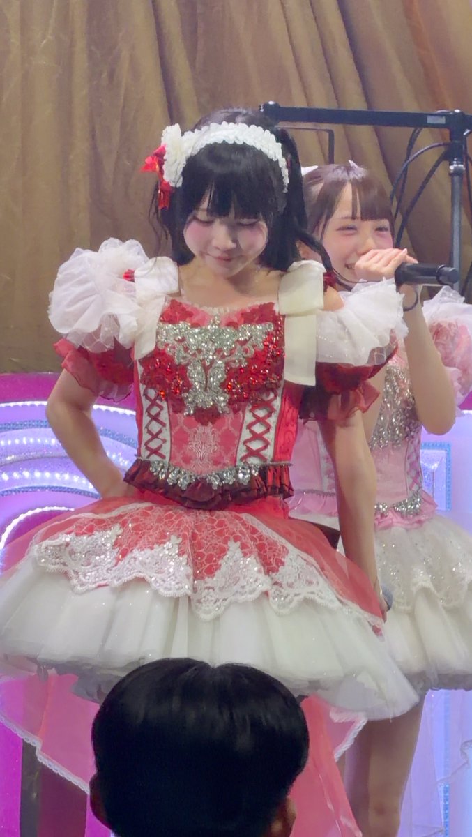 Two performers in elaborate red and white frilly dresses with sequined bodices are on stage; the foreground performer wears a white lace headband with a red bow, while the background performer holds a microphone and laughs.