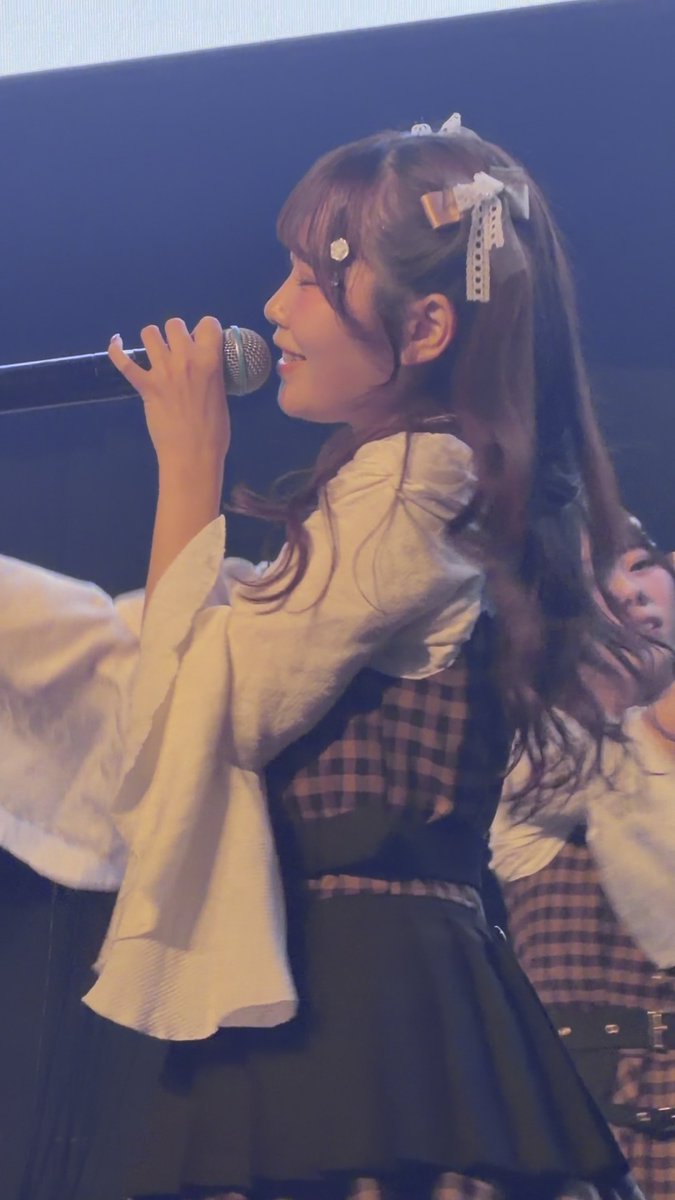 A young woman with long dark hair sings into a microphone on stage, wearing a checkered outfit with puffy white sleeves and decorative hair ribbons.