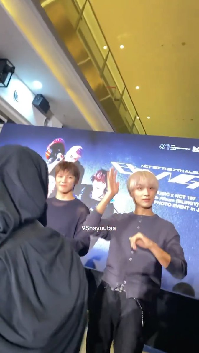 Two young men with blonde and dark hair on stage at an NCT 127 fan event, with one waving and a person in a dark hooded jacket in the foreground.