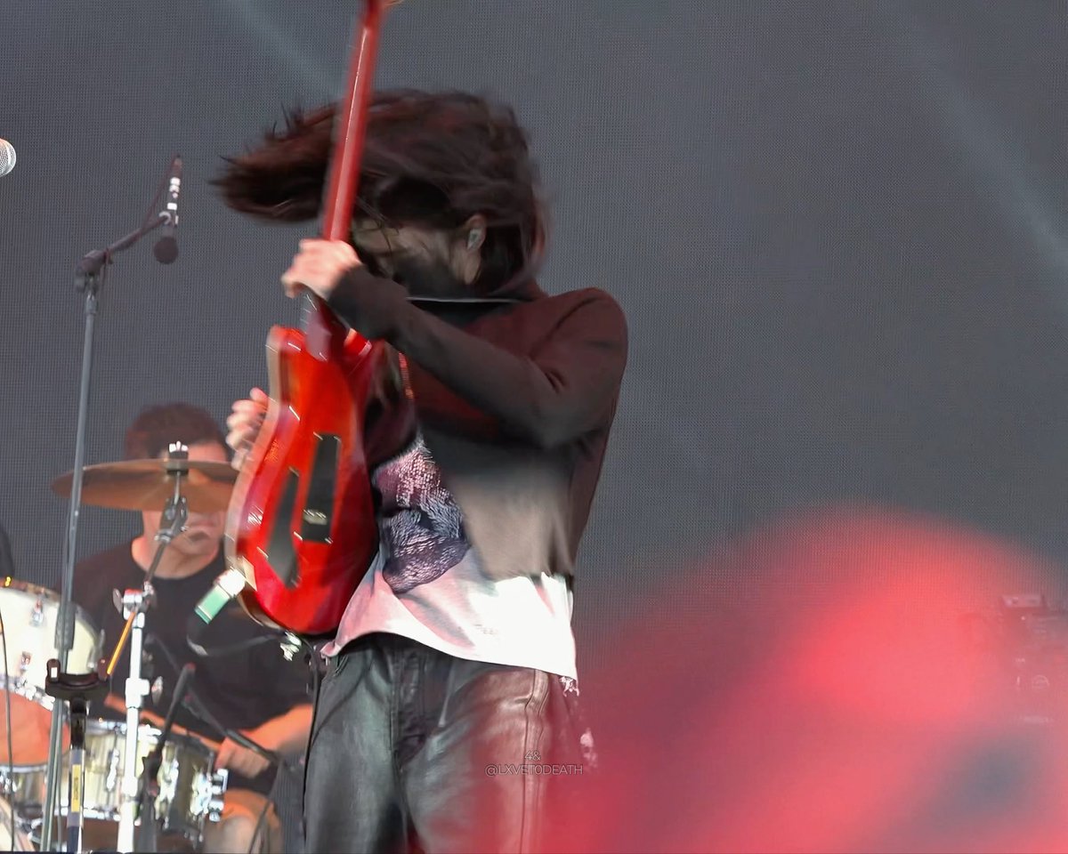 Concert photo of a musician energetically headbanging while playing guitar, captured with motion blur. The image is credited to '@LXVE.TODEATH'.