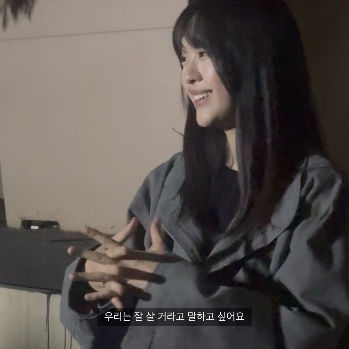 A young Korean woman with long dark hair, wearing a gray coat, sits and gestures with her hands while smiling during a conversation.
