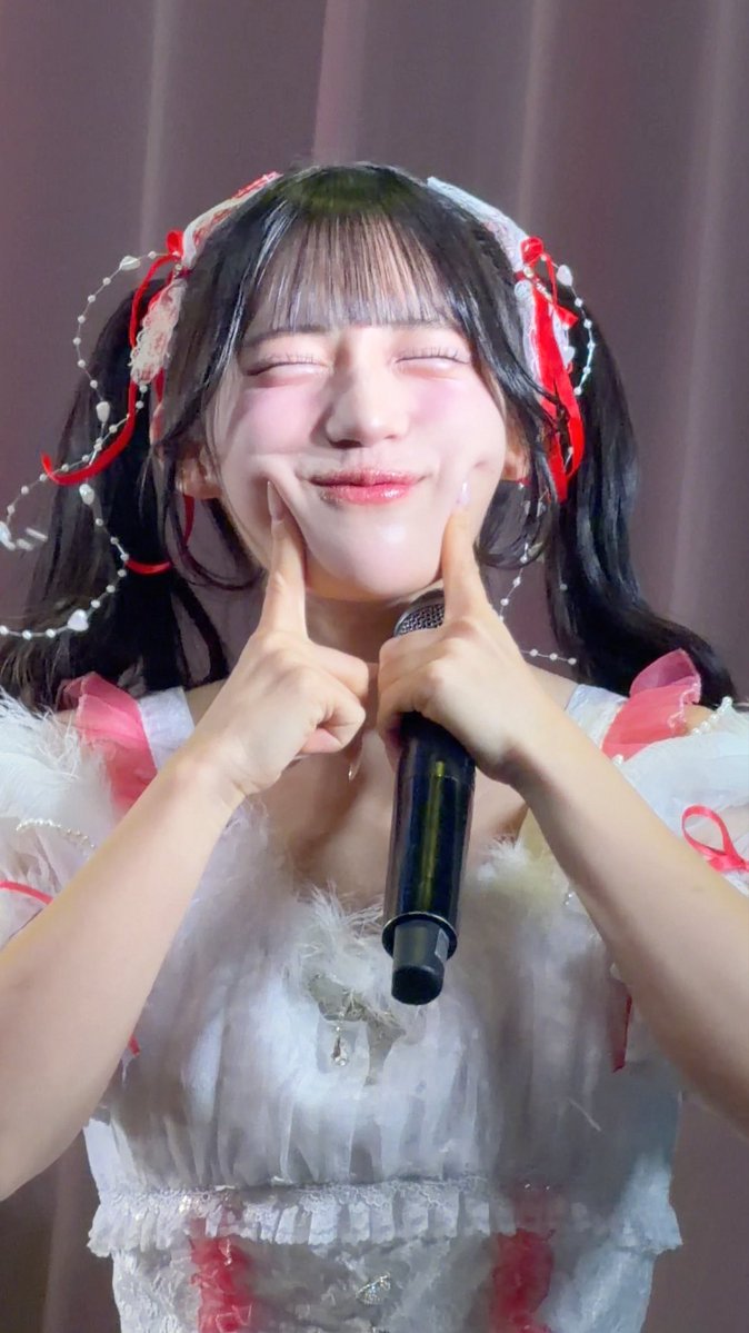 A young performer with dark hair in twin tails and a frilly white outfit poses with her eyes closed, smiling while pointing to her cheeks and holding a microphone.