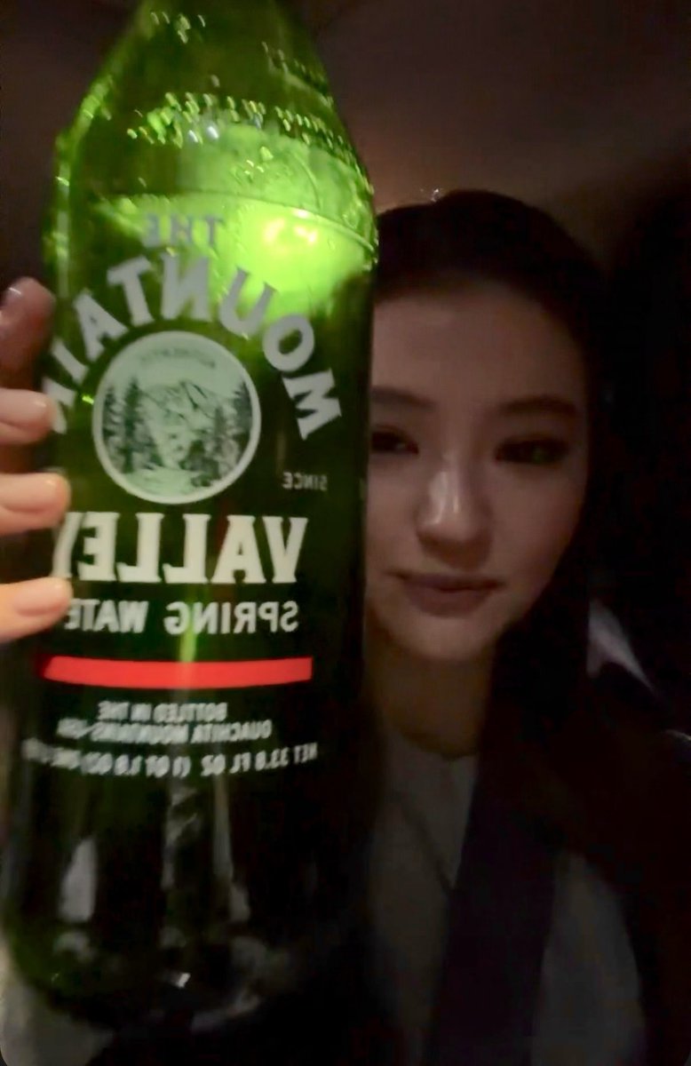 A young woman smiles while holding up a large green bottle of Mountain Valley Spring Water, with the label appearing reversed.
