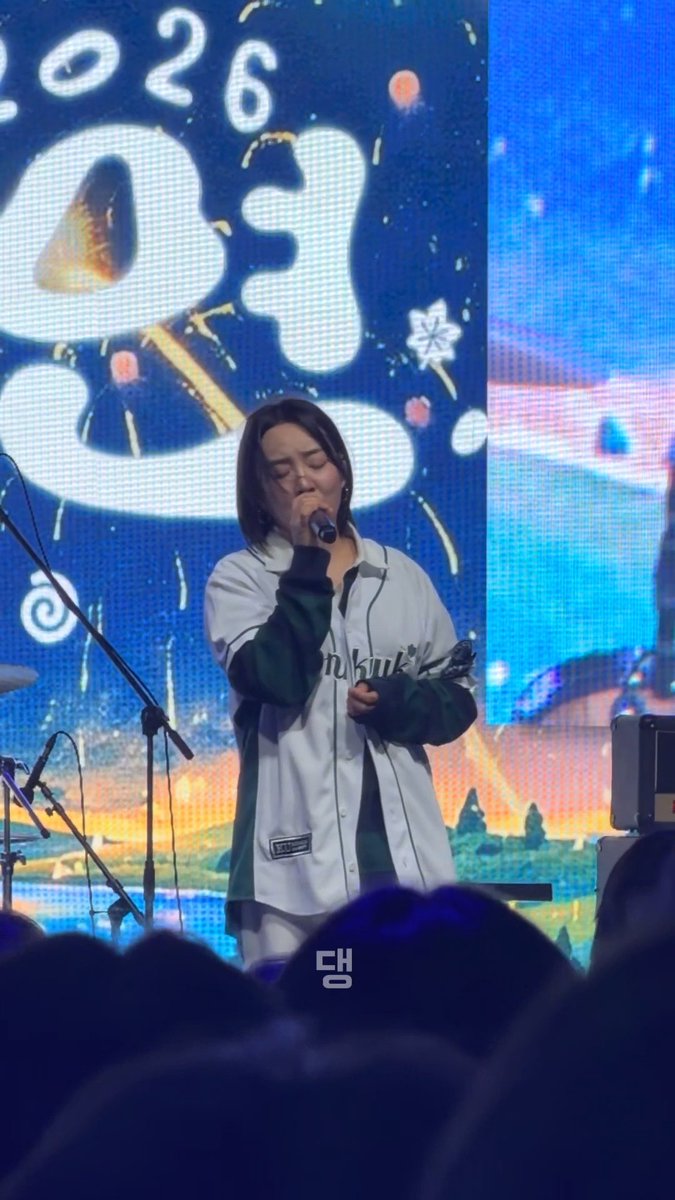 A performer with shoulder-length dark hair sings into a microphone on stage, wearing a green and white varsity-style jacket with 'Onhuk' visible on the front, in front of a screen displaying '2026' and Korean text.