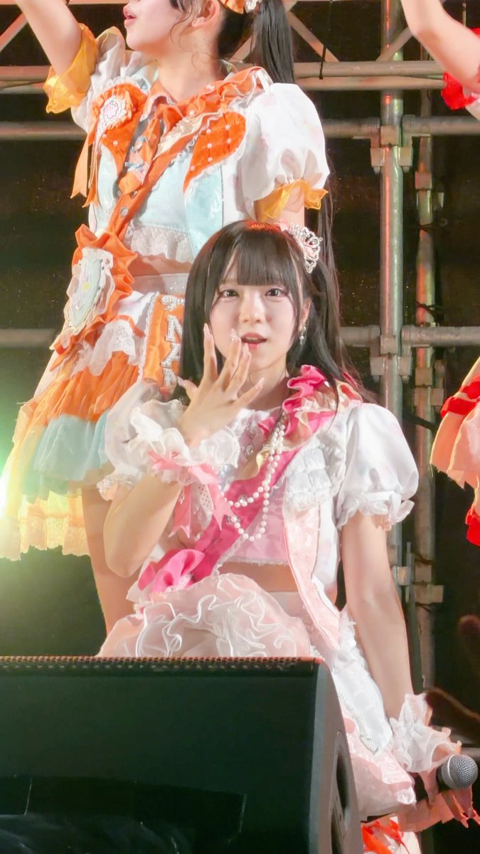 A young woman in a frilly pink and white idol costume stands on stage holding a microphone, with another performer visible behind her.