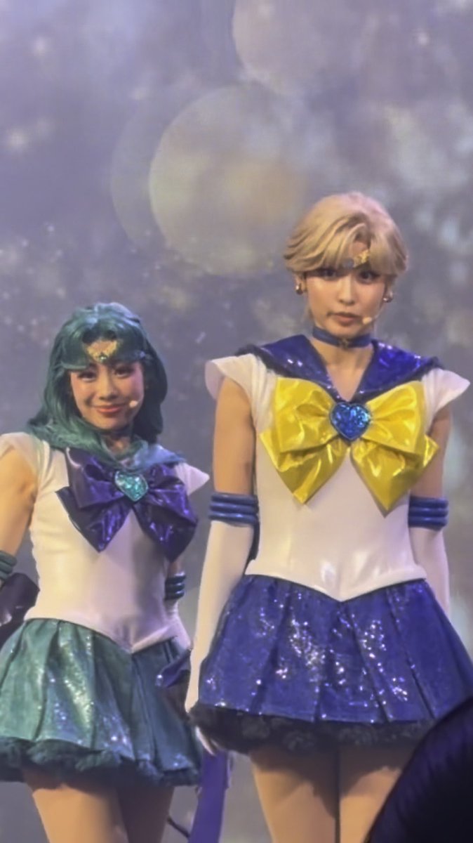 Two performers in Sailor Moon musical costumes, dressed as Sailor Neptune (teal) and Sailor Uranus (blue) with blonde hair, standing on a stage with a starry backdrop.