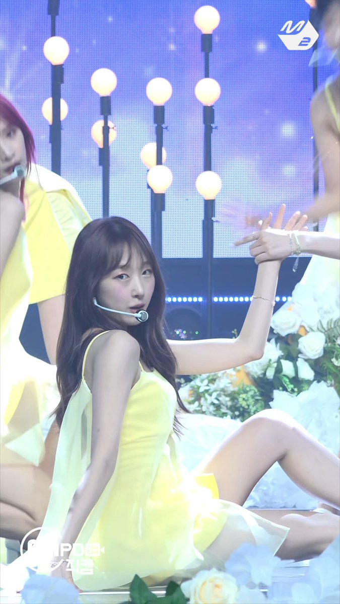 K-pop performance featuring a young woman in a yellow dress with a headset microphone, performing on stage with floral decorations and illuminated spherical lights in the background.