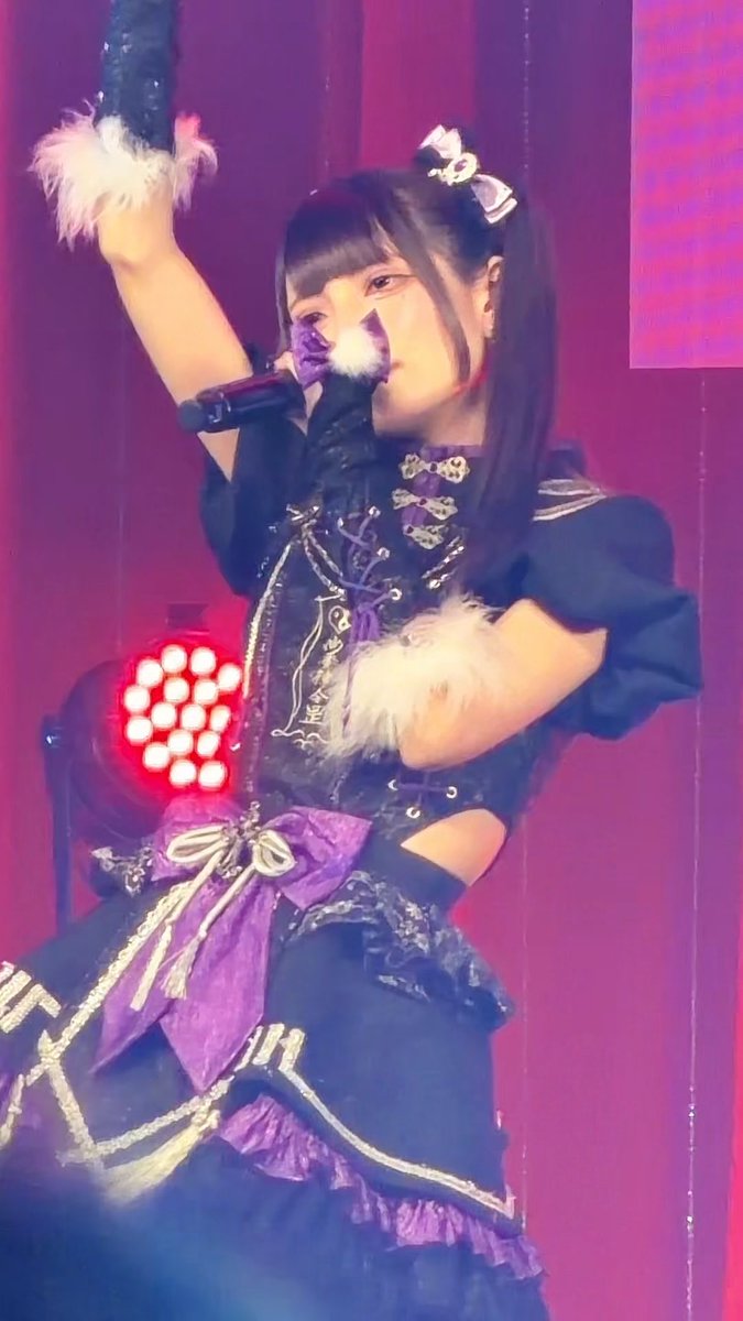 A performer in an elaborate black and purple gothic-style costume with feathered cuffs and decorative chains sings into a microphone on stage under pink lighting.