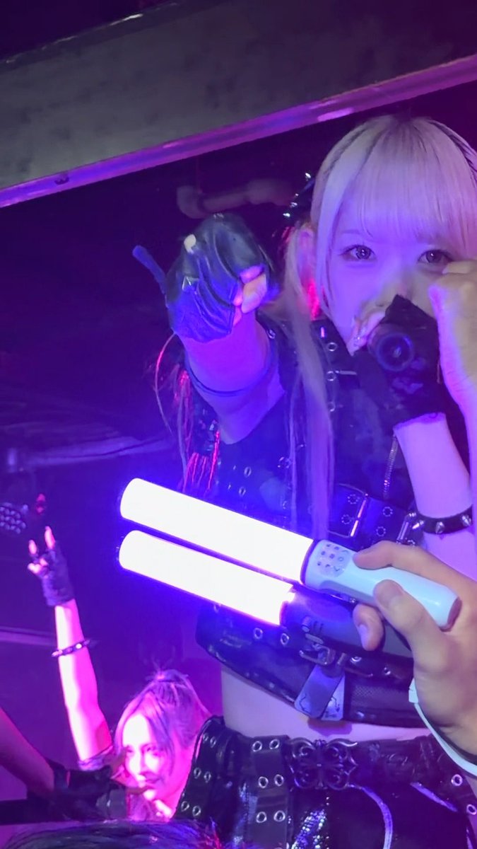 A performer with blonde hair, wearing a black leather harness outfit and black gloves, holds a glowing double light stick while taking a selfie in a purple-lit venue.