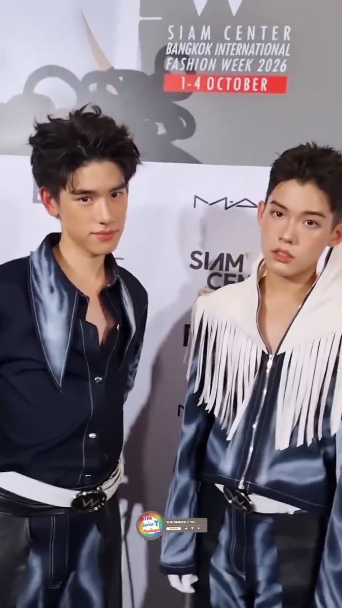 Two young men pose in coordinated dark blue satin outfits at Siam Center's Bangkok International Fashion Week 2026 backdrop, with the left model wearing a collared shirt and the right model sporting a fringed jacket. Visible text on the backdrop promotes "SIAM CENTER BANGKOK INTERNATIONAL FASHION WEEK 2026" from "1-4 OCTOBER," aligning with the post identifying the duo as Yugeen and Yugene at the event.