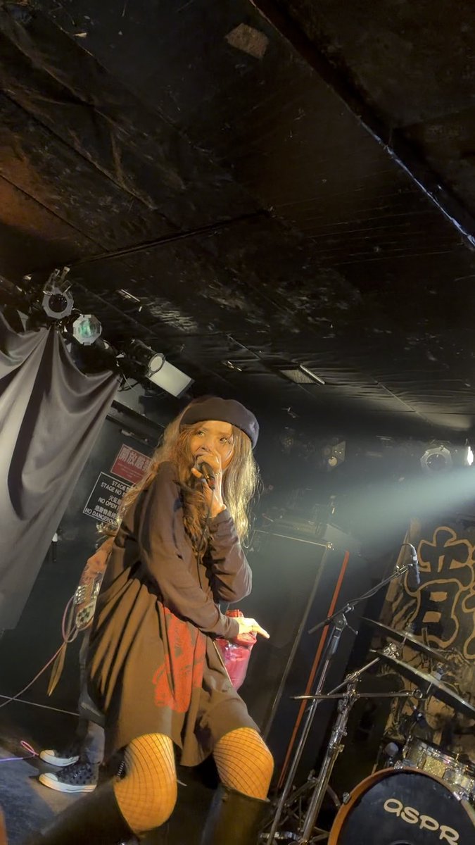 A female performer with long hair and a black beret sings into a microphone on a dimly lit stage, wearing a dark dress with red details and yellow fishnet stockings, with an 'aspra' drum kit visible nearby.