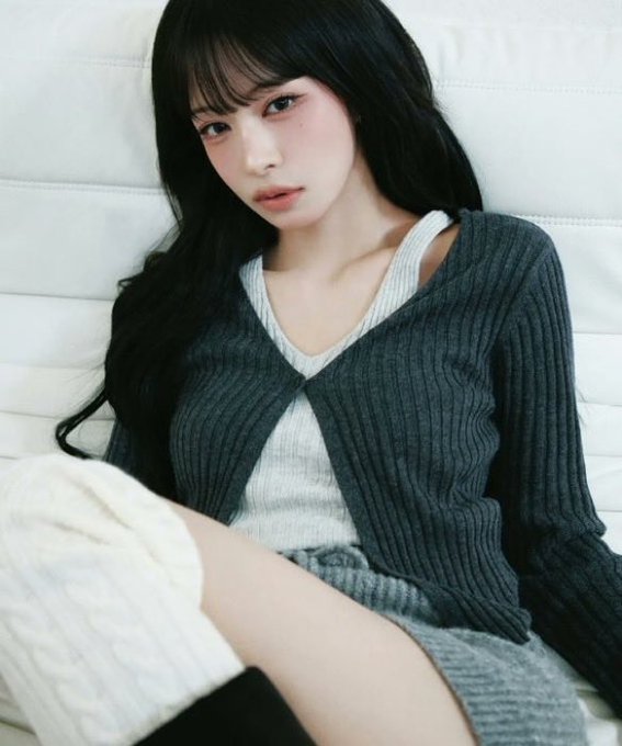 A soft-toned portrait of a young woman with long dark wavy hair and bangs, wearing a dark gray ribbed cardigan over a light top, seated against a tufted white headboard.