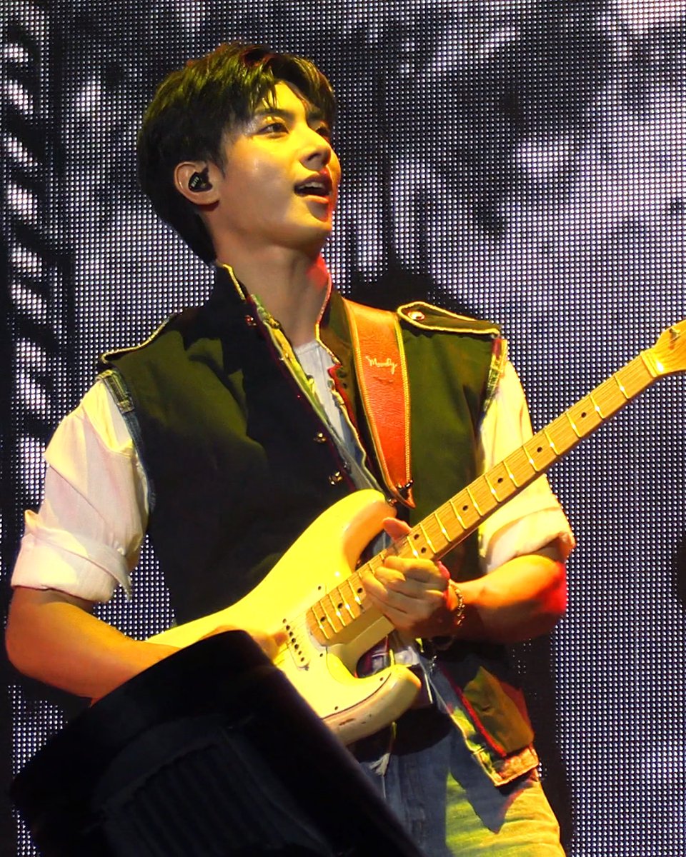 A male guitarist performs on stage with an electric guitar, wearing a green vest over a white shirt and an in-ear monitor, illuminated by a bright LED screen backdrop.