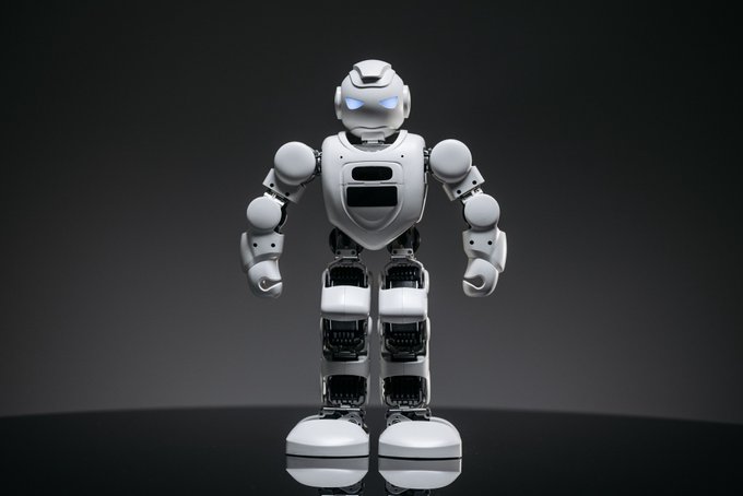 A white humanoid robot with glowing blue eyes stands against a dark gradient background, featuring articulated joints and a chest panel with a dark screen.