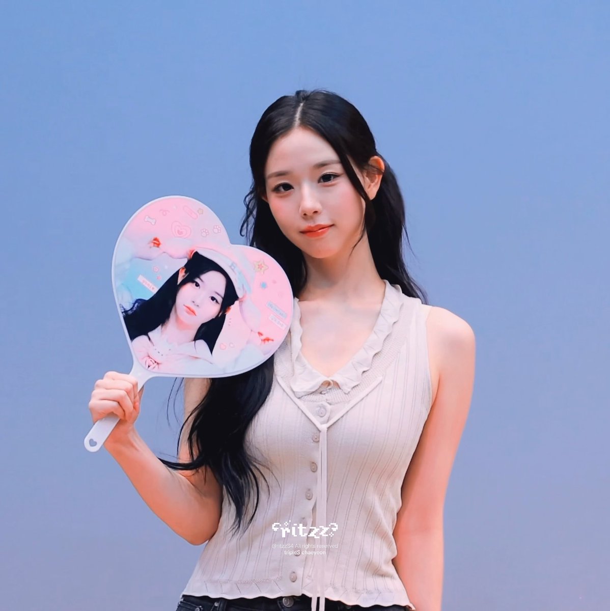 A young woman with long dark hair holds a heart-shaped fan with a pink illustration, wearing a cream-colored sleeveless top against a blue gradient background; text reads 'ritze' and 'tripleS haeyeon'.