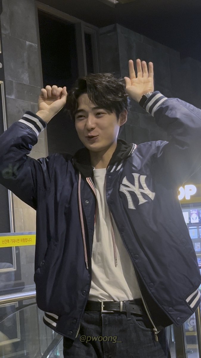 A young man wearing a navy blue New York Yankees varsity jacket over a white tee, posing with both hands raised playfully near his head in an indoor setting.