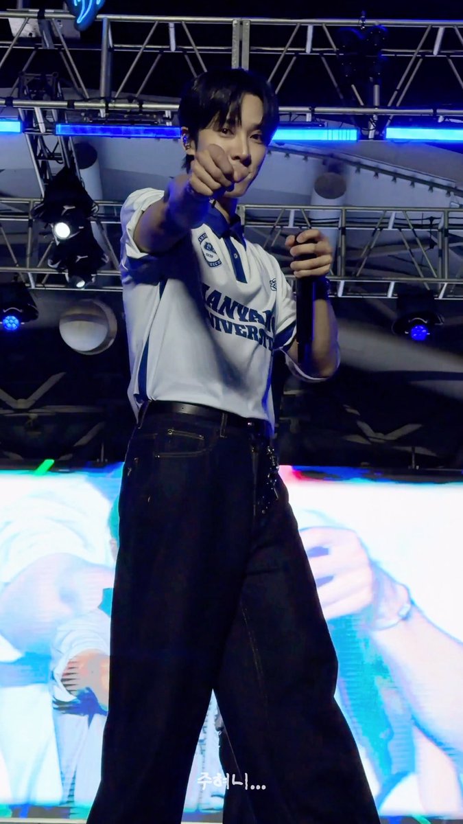 A man on stage in a white 'YONSEI UNIVERSITY' polo shirt and black pants, pointing at the camera while holding a microphone, with Korean text '주혜니...' overlaid at the bottom.