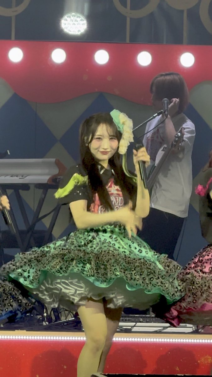 A female performer in a black, yellow, and green dress holds a microphone on a stage, with a guitarist and a keyboard visible in the background.