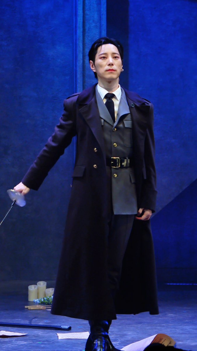 A performer on stage wears a long dark trench coat over a gray suit, holding a long thin item, appearing to be part of a theatrical production.