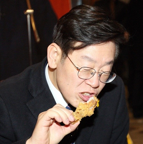 A man wearing glasses and a dark suit is eating a piece of fried chicken.