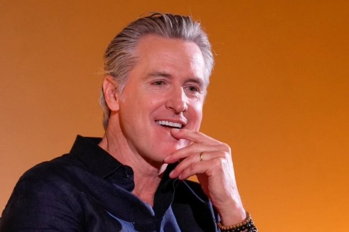 A portrait of California Governor Gavin Newsom smiling with his hand on his chin, wearing a dark shirt and a beaded bracelet, against an orange background.