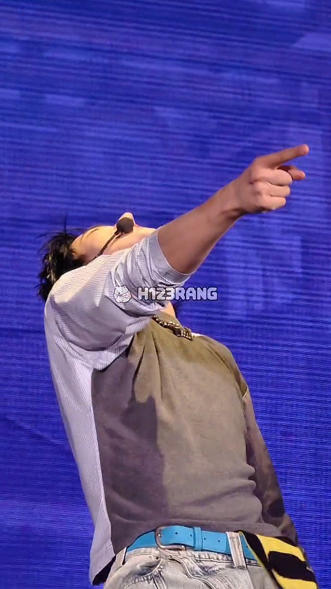 A performer wearing a light striped shirt, dark vest, turquoise belt, and jeans points dramatically upward against a blue background, with a watermark reading 'H123RANG'.