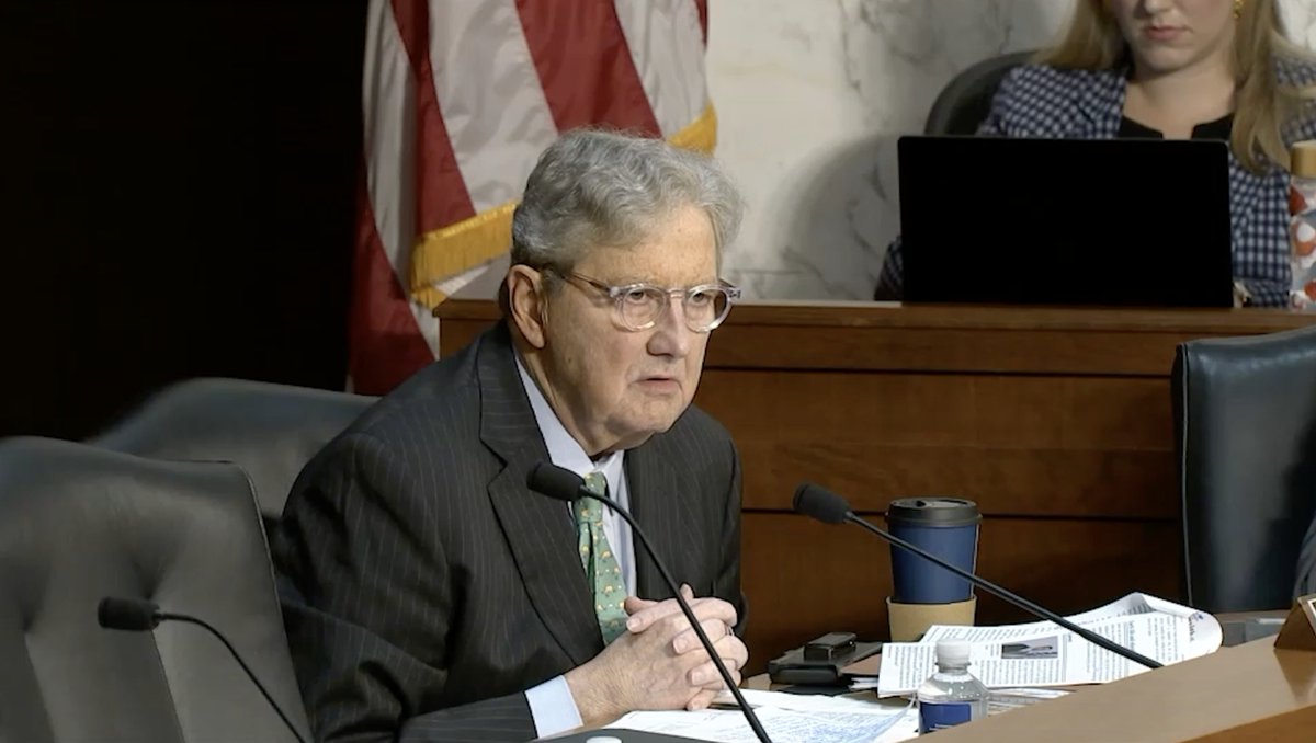 An older man with glasses, wearing a pinstripe suit and green tie, sits at a wooden desk with microphones, papers, and a blue tumbler, appearing to speak during what looks like a Senate hearing.