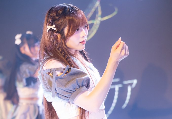 A young woman with braided pigtails and a star hair clip performing on stage in a frilly outfit, with another performer visible in the background.