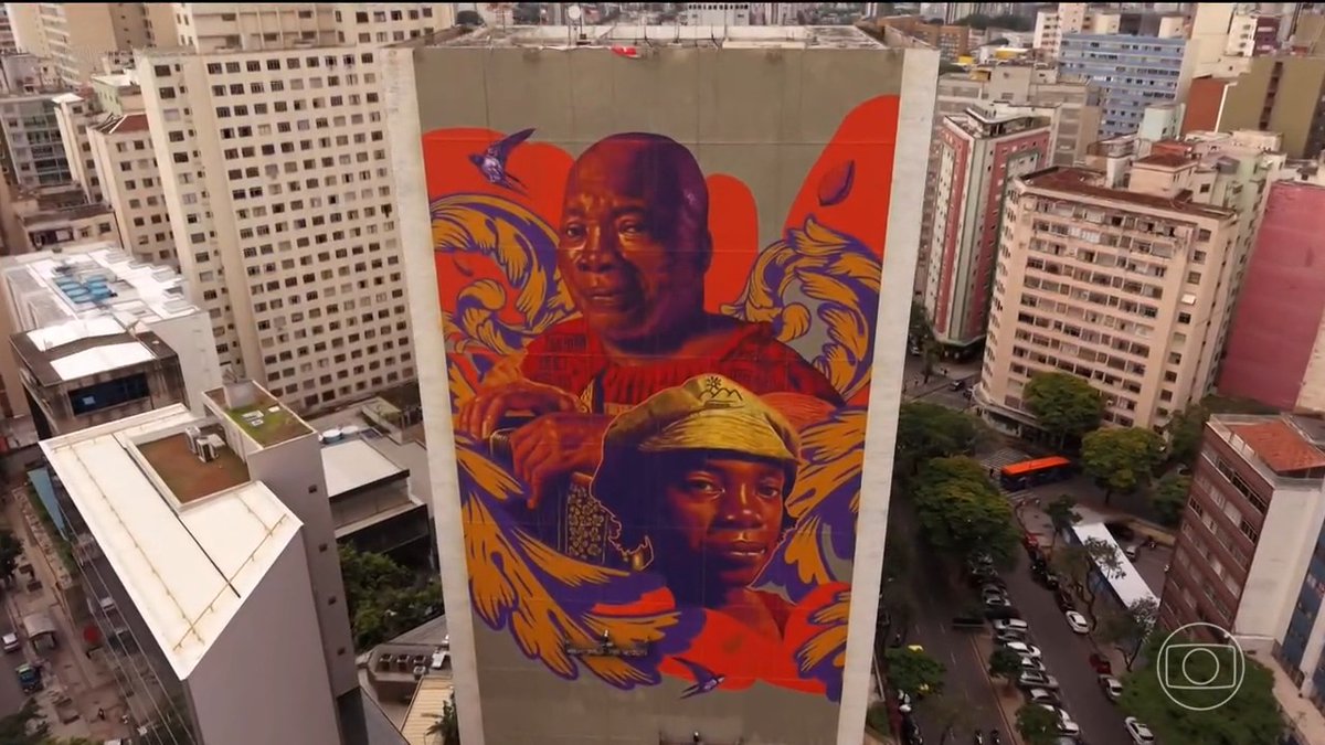 An aerial view of a large building mural depicting the faces of a Black woman and child surrounded by purple birds and orange floral patterns, set against a city skyline.