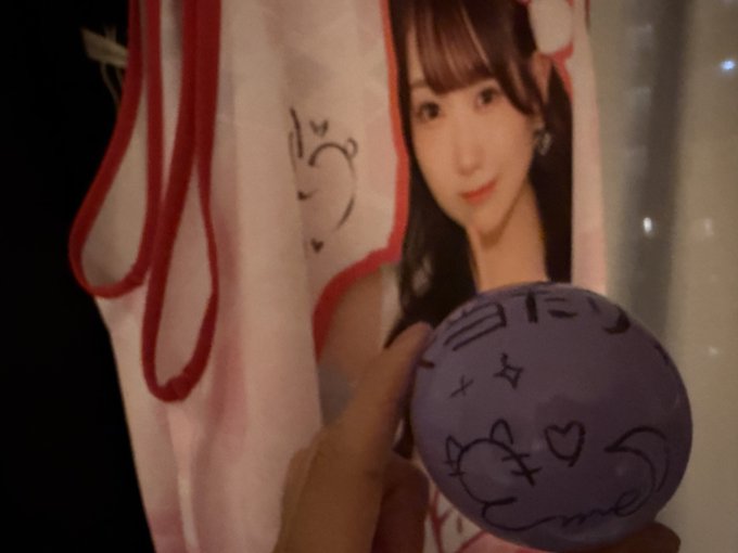 A fan holds up a signed purple plush ball featuring cute doodles like a bear face, hearts, and stars, alongside an acrylic stand or towel of what appears to be the Japanese idol Yuki (as referenced in the caption about catching/collecting her merchandise).