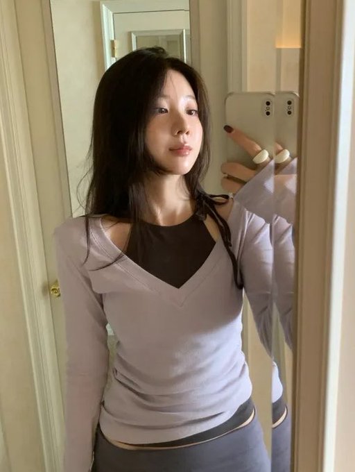A woman with long dark hair takes a mirror selfie while wearing a layered long-sleeve top featuring a light purple outer layer with a deep V-neck cutout over a black underlayer. The caption references a reward link for new YUUNI apparel, highlighting the outfit's stylish design.