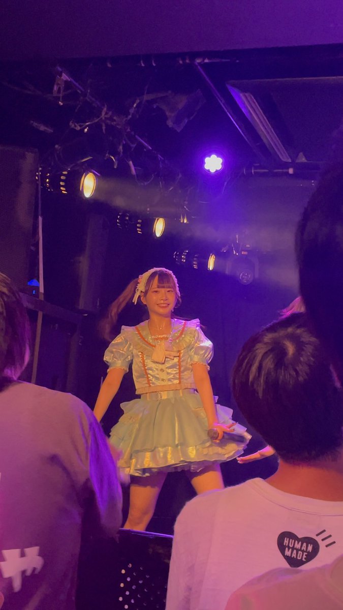 A performer in a light blue frilly idol dress with twin-tails and a bow stands on stage holding a microphone, illuminated by purple and yellow stage lights, while audience members watch from below—one wearing a white "HUMAN MADE" shirt. The image captures a small live venue idol performance, matching the post's regret about not bringing a better camera to photograph the performer (Miku).