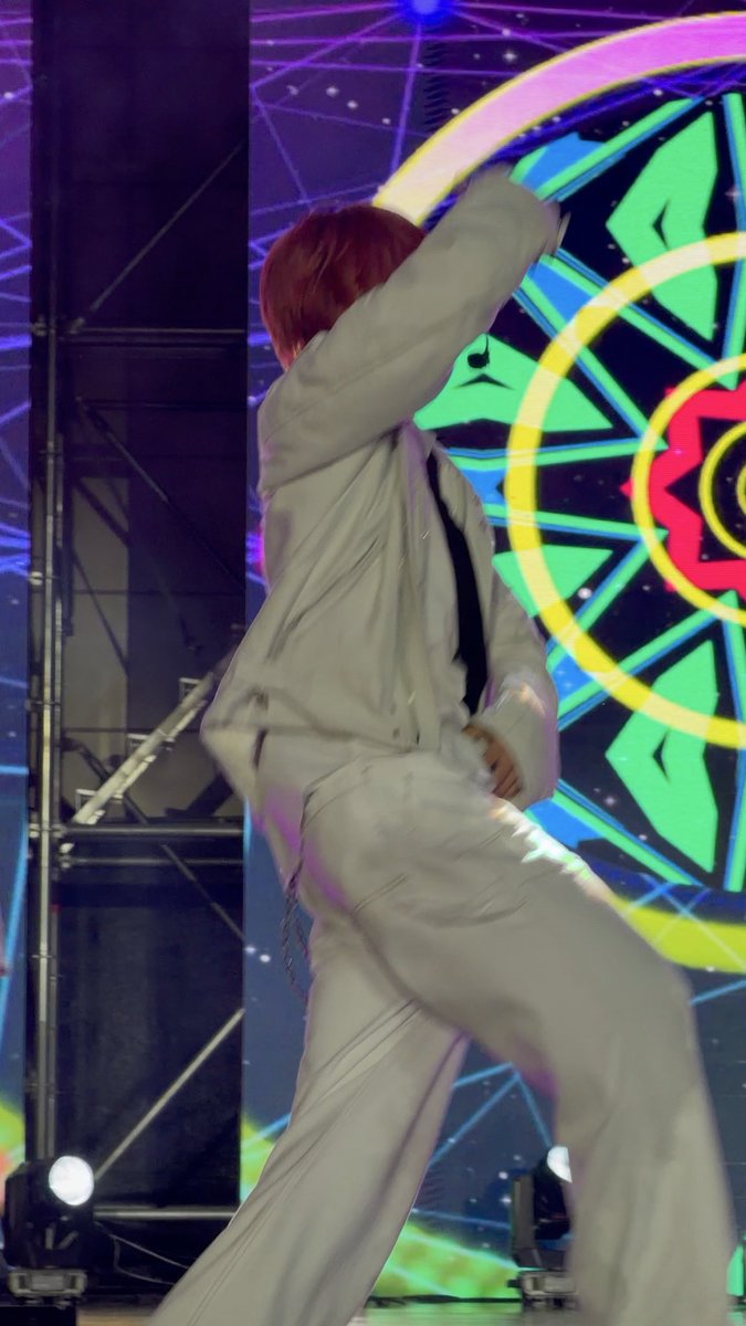 A performer with reddish hair in a white suit and pants dancing energetically on stage, with one arm over their face, against a backdrop of a colorful circular stained-glass-like design.