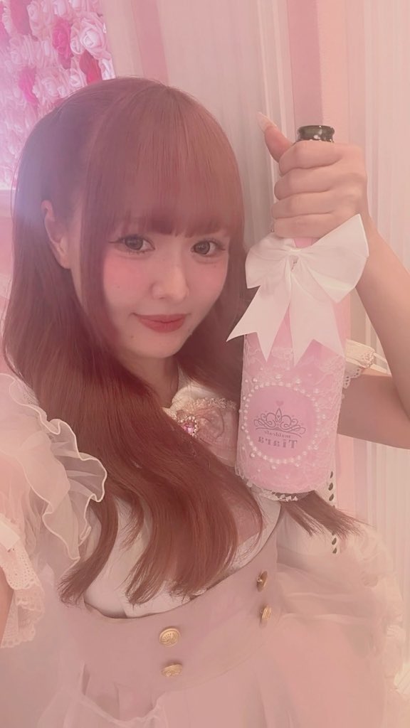 A young woman with reddish-brown hair and bangs wearing a pink outfit, holding up a pink bottle with a white bow in a pink-decorated setting.