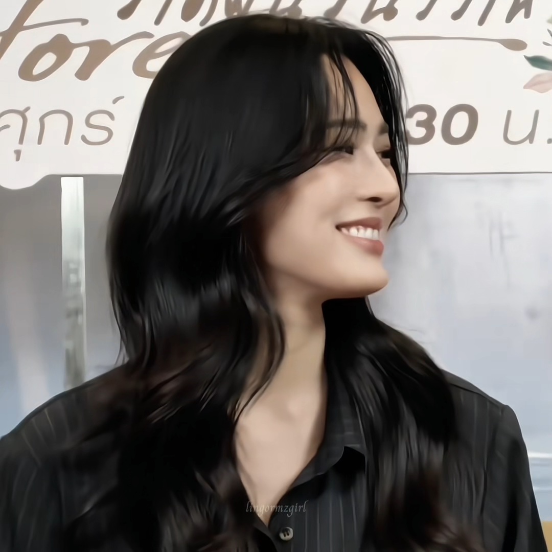 A close-up profile photo of a smiling woman with long, wavy black hair wearing a black shirt, with Thai text and a watermark 'lunormzgirl' visible in the background and foreground.