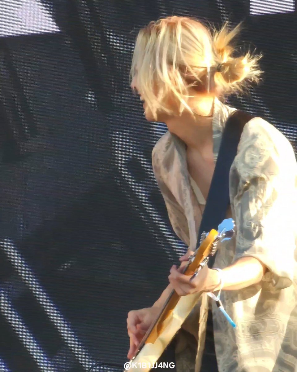 A blonde musician with hair pulled into a messy bun plays an electric guitar while performing on stage against a dark backdrop, with a watermark reading '@K1B1J4NG' visible at the bottom.