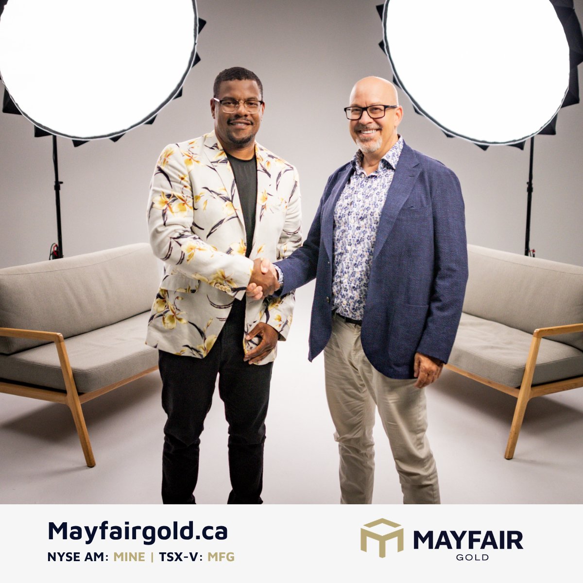 A promotional photo of two men shaking hands in a studio setting, advertising Mayfair Gold (Mayfairgold.ca) with ticker symbols NYSE AM: MINE and TSX-V: MFG.