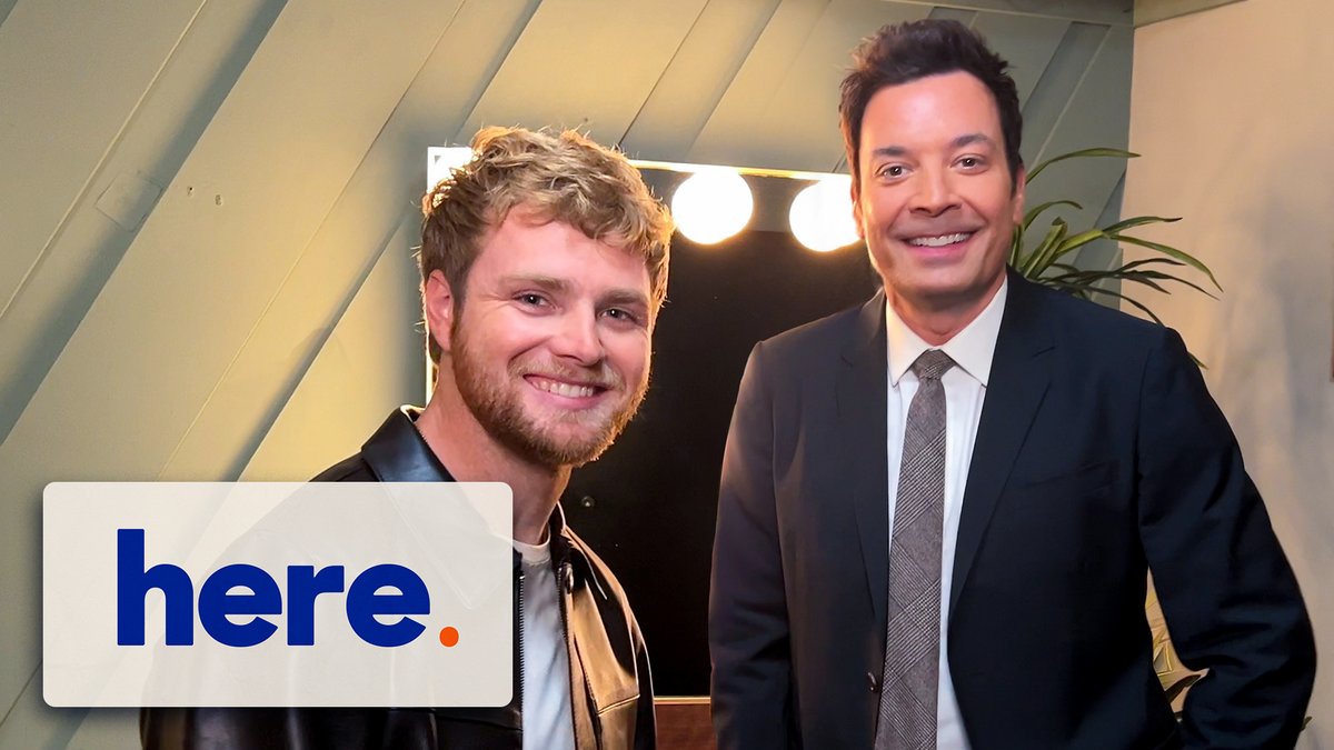 Singer Alex Warren stands smiling beside Jimmy Fallon in a studio setting, with a "here." logo (likely AEW's *Here Comes the Sun* branding or similar) overlaid in the corner of the photo.